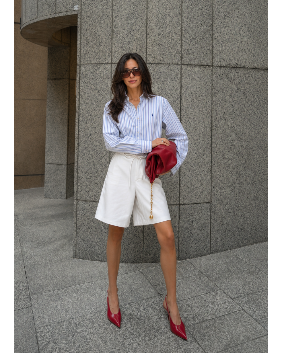Woman in a striped shirt and white shorts holding a red clutch against a gray building.