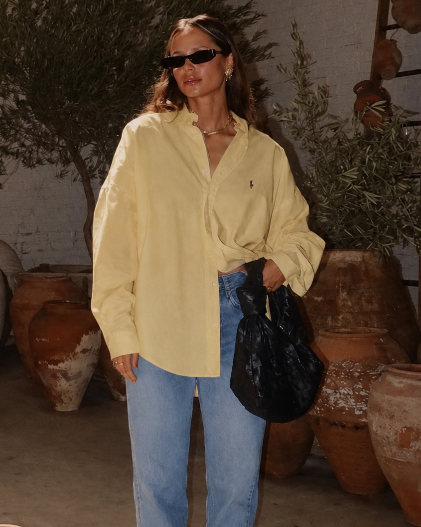 Woman in a yellow shirt and blue jeans holding a black bag in an outdoor setting with potted plants.
