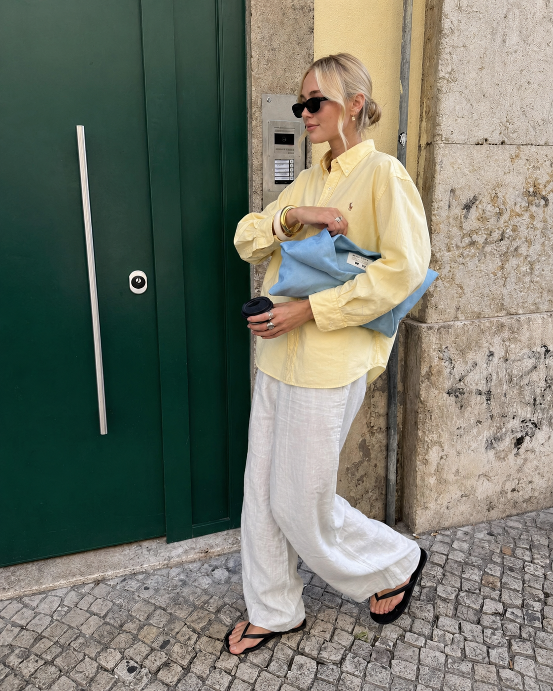 Woman in a yellow shirt and white pants holding a blue clutch against a textured wall and green door.