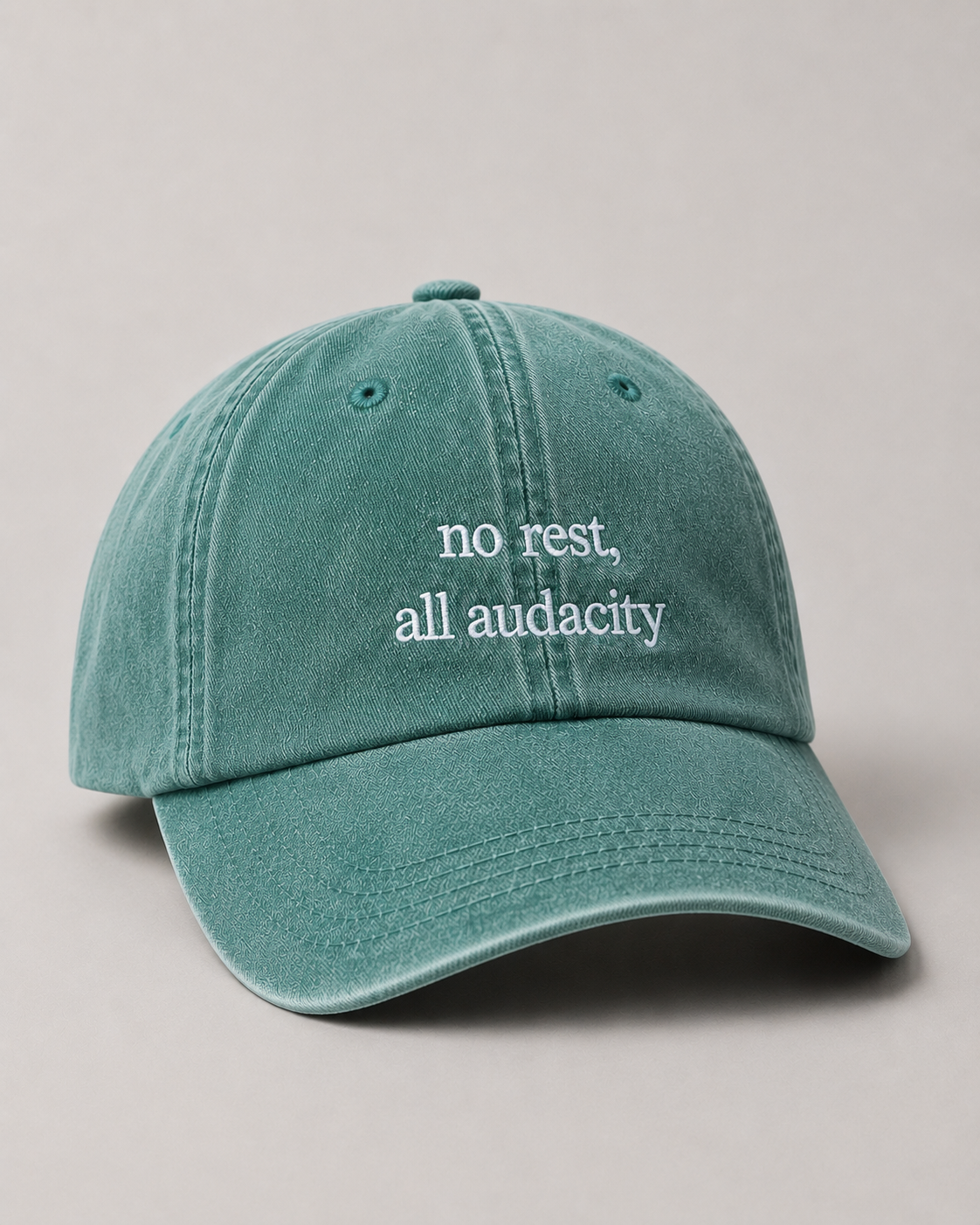 Teal cap with 'no rest, all audacity' text on a beige background
