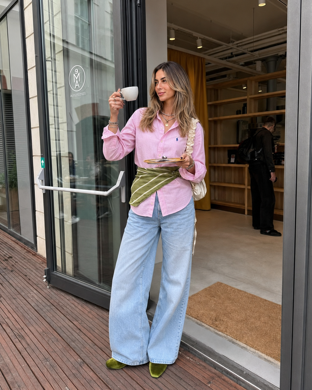 Woman in pink shirt and light blue jeans holding a coffee cup outside a building.