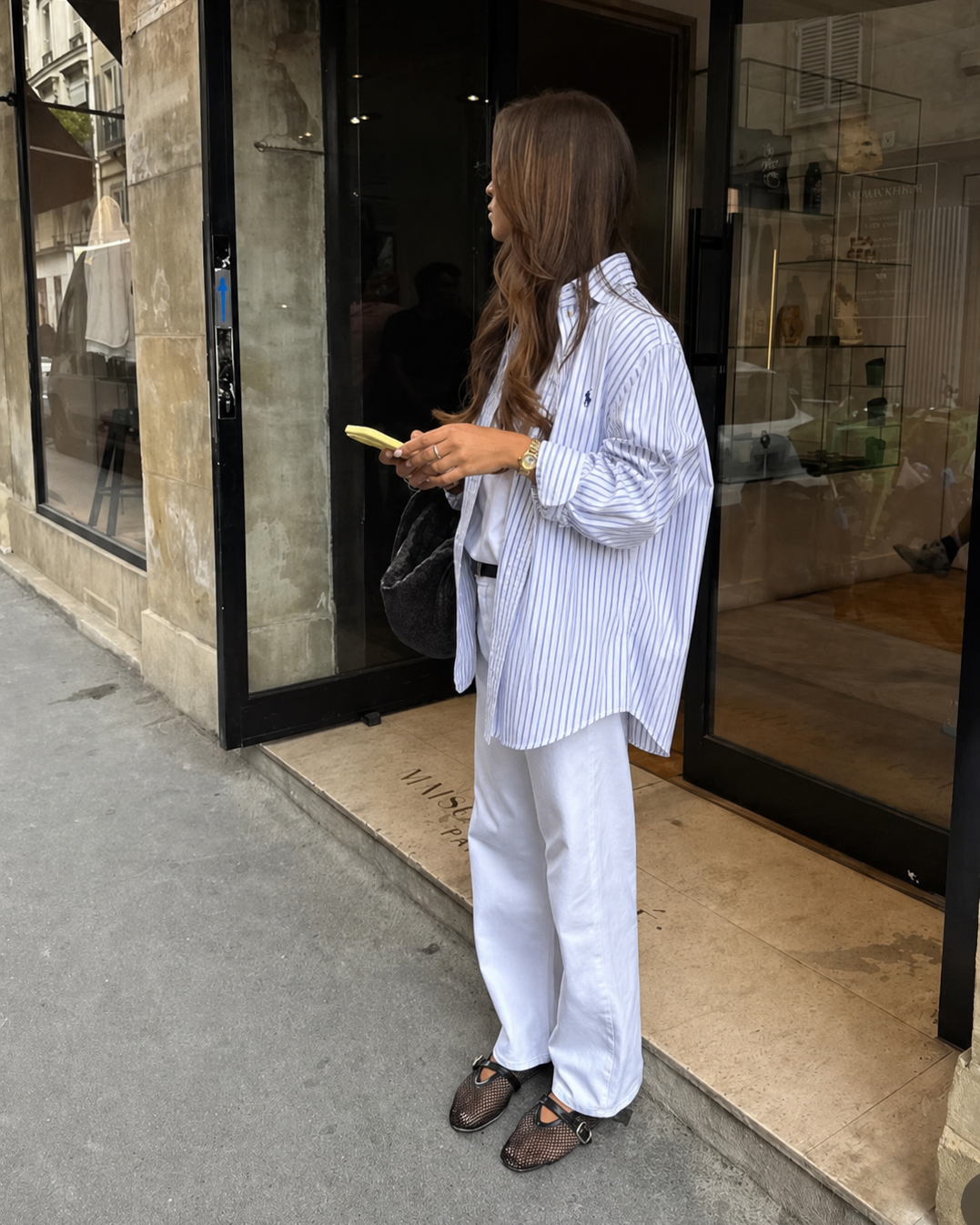 Person standing outside a store, holding a phone and wearing a striped shirt and white pants.