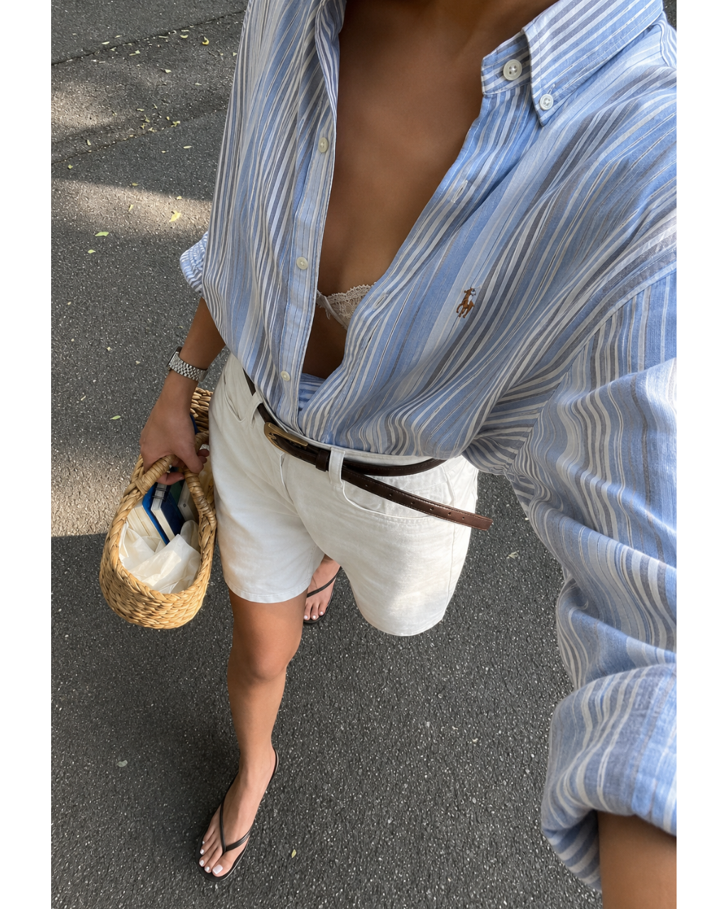 Person wearing a blue striped shirt and white shorts, holding a woven bag on a street.