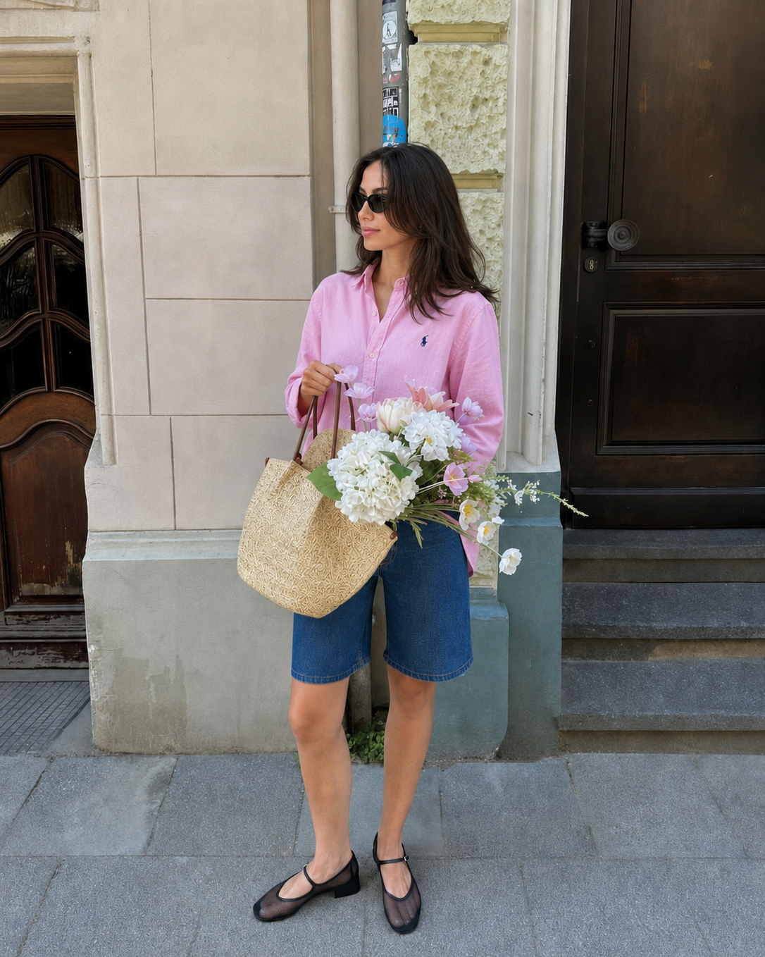 Woman in pink shirt and blue shorts holding a straw bag and flowers, standing on a sidewalk.
