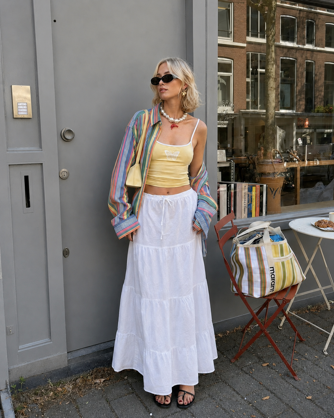 Woman in colorful striped jacket, yellow top, and white skirt standing outdoors with a building in the background.