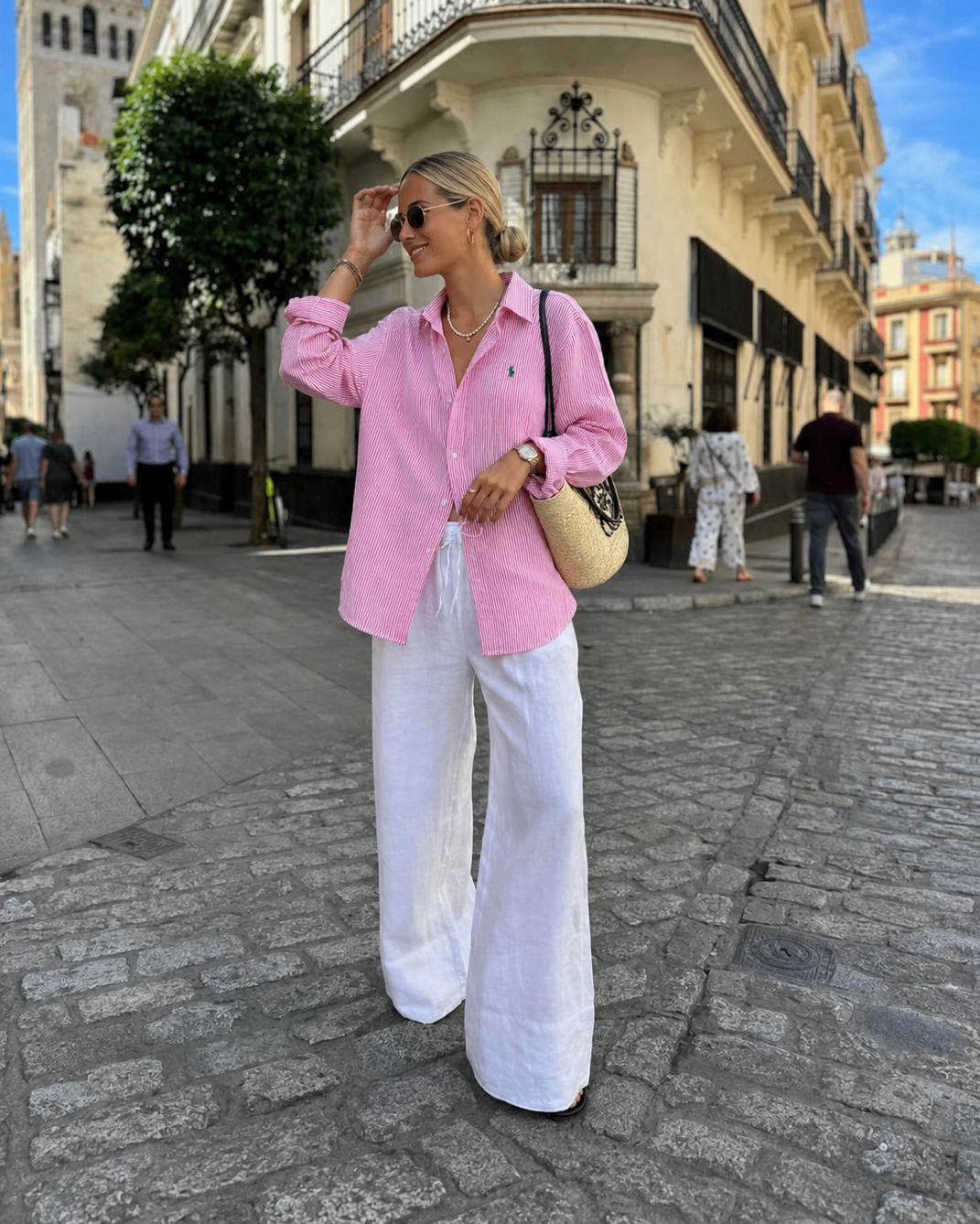 Woman in pink shirt and white pants walking on a street with buildings in the background