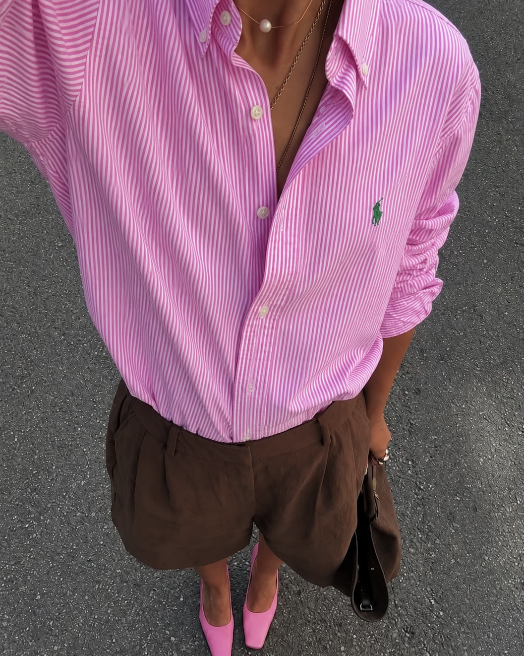 Person wearing a pink striped shirt with a logo, brown shorts, and pink shoes on a gray pavement.